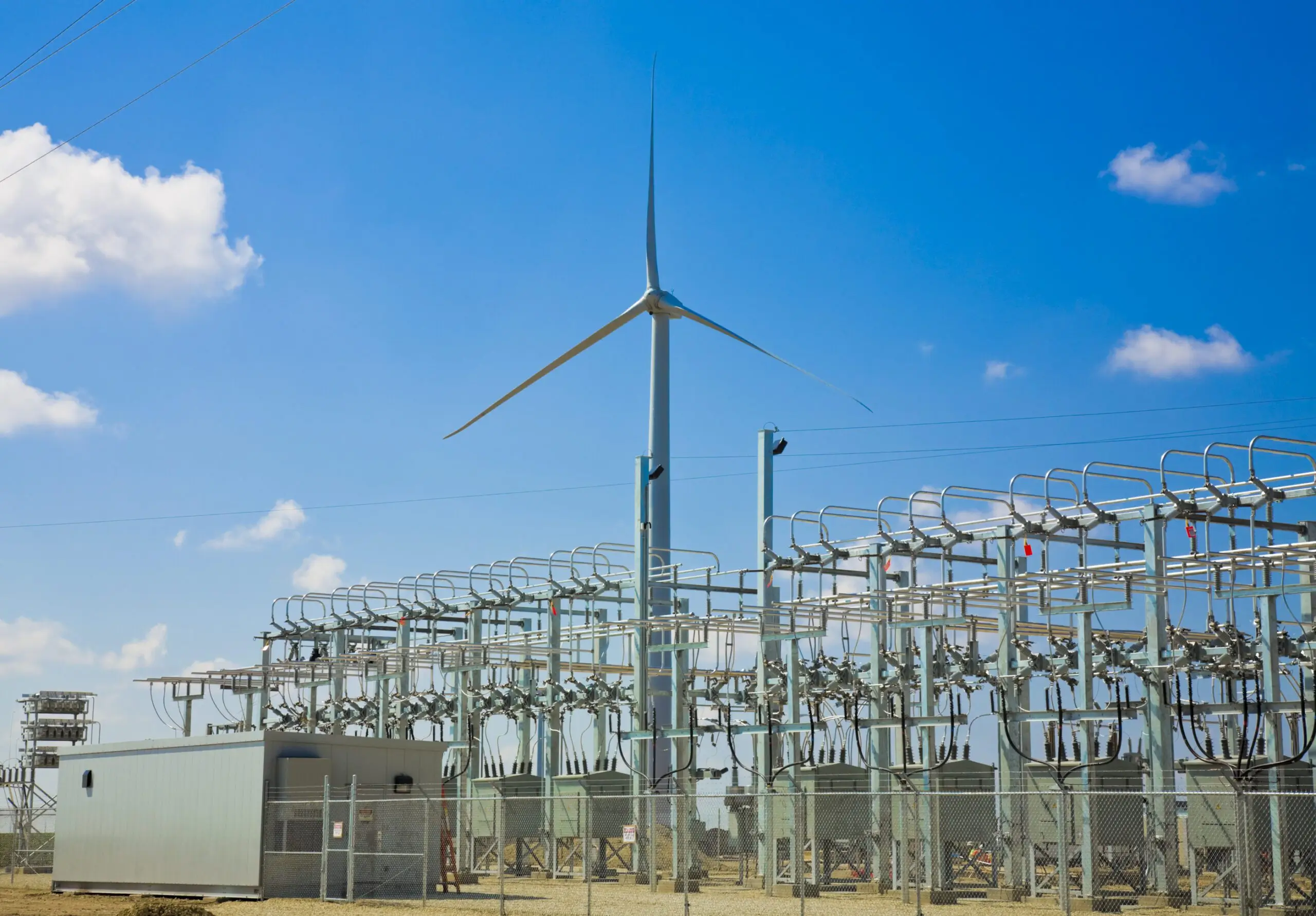 A wind turbine standing tall behind a power substation with various electrical components, under a clear blue sky, representing renewable energy and electrical infrastructure.